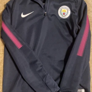 Manchester City långärmad träningströja Nike - Mörkblå långärmad Manchester City träningströja från Nike med halv dragkedja och klubbmärke på bröstet. Snygga rosa detaljer längs ärmarna och tillverkad i ett lätt, sportigt material som andas. Perfekt för dig som älskar fotboll och vill sticka ut på planen.