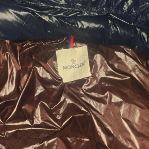Mörkblå Moncler pufferjacka med huva - Snygg mörkblå pufferjacka från Moncler med glansig finish, huva och dragkedja med logodetaljer. Jackan har brunt innerfoder och klassisk Moncler-patch på ärmen. Perfekt för kalla dagar och riktigt stilren look. SIZE XL Funkar bra för Folk med L också