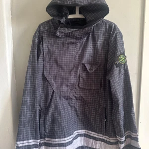 Grå rutmönstrad vindjacka Stone Island - Snygg grå vindjacka från Stone Island med vitt rutmönster och stor logga på ryggen. Jackan har huva, bröstficka med lock och klassisk patch på ärmen. Perfekt för dig som vill sticka ut med streetwear-vibe.