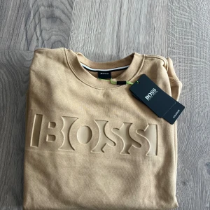 Beige sweatshirt från BOSS - Snygg beige sweatshirt från BOSS med upphöjt logotyptryck framtill. Tröjan har rund halsringning och långa ärmar, perfekt för en clean och stilren look. Tillverkad i mjuk bomull som känns skön mot huden.