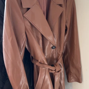 Brun skinn trenchcoat med bälte - Snygg brun trenchcoat i skinnimitation med breda slag, knappar framtill och matchande bälte i midjan. Jackan har en klassisk, rak passform och långa ärmar. Perfekt för dig som vill ha en trendig och stilren look.