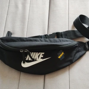 Svart Nike magväska med AIK-logga - Snygg svart magväska från Nike med vit logga och AIK-emblem på framsidan. Väskan har justerbart band, dubbla dragkedjor och är tillverkad i slitstark polyester. Perfekt för att bära mobilen, nycklar och andra småsaker när du är på språng.