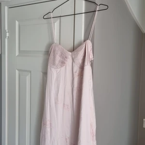 Ljusrosa långklänning från Adoore - Supersöt ljusrosa långklänning från Adoore med tunna axelband och plisserad byst. Klänningen har subtila blommiga detaljer och en lätt utsvängd nederdel. Perfekt för dig som vill ha en romantisk och feminin look. Använd 1 gång