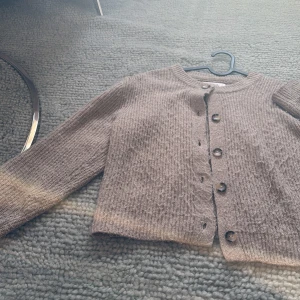 Beige stickad kofta från Zara - Mysig beige stickad kofta från Zara med rund halsringning och stora knappar framtill. Koftan har långärmad passform och är perfekt att slänga över en t-shirt eller linne. Den ribbade strukturen ger en trendig och avslappnad vibe.