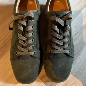 Mörkgröna mocka sneakers från Louboutin - Snygga mörkgröna sneakers från Louboutin i mjuk mocka med rund tå och platt sula. Skorna har matchande gröna snören och broderad logga på hälen. Insidan har en vit sula med präglad Paul Smith-signatur. Perfekt för dig som gillar stilrena detaljer.