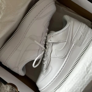 Nike Air Force 1 Shadow vit sneakers - Nike Air Force 1 Shadow i helvit färg, storlek 40,5 . Helt nya i kartong, säljes då jag har problem med ryggen och endast använder ett märke på skor. Skickas i original kartong
