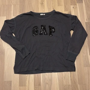 Svart långärmad tröja GAP XS - Svart långärmad tröja från GAP i bomull med glittriga paljetter på bröstet som bildar loggan. Avslappnad passform och rund halsringning, perfekt för en chill look.