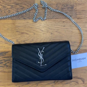 Svart quiltad axelväska från YSL - Snygg svart axelväska från YSL i quiltat skinn med silverfärgad kedja och ikonisk YSL-logga framtill. Väskan har ett rött innerfoder, dragkedjefack och magnetlås. Perfekt storlek för det viktigaste och ger en lyxig vibe till din outfit.