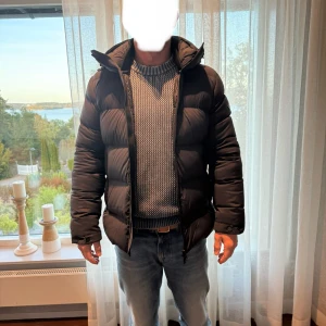 Svart pufferjacka från J.Lindeberg - Säljer en svart pufferjacka från J.Lindeberg med huva och dragkedja. Jackan är vadderad och har en stilren look med diskret logga på ärmen. Perfekt för kalla dagar och passar dig som gillar en modern och enkel stil.