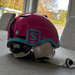 Rosa Salomon hjälm Kid M - Cool rosa hjälm från Salomon med turkos detalj runt nederkanten och vitt meshfoder. Hjälmen har justerbart hakband och öronskydd i vitt mesh. Perfekt för vinterns äventyr i backen eller på isen.