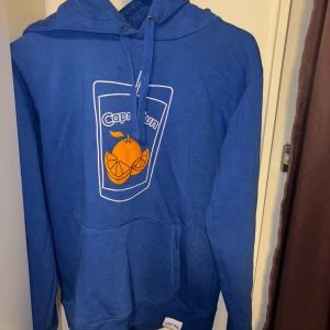 Blå Capri-Sun hoodie med tryck - Blå hoodie från Capri-Sun med coolt tryck av ett glas och apelsiner på bröstet. Tröjan har huva med snörning och en stor magficka. Perfekt för dig som gillar färg och vill sticka ut lite extra. Har små fläckar som visas på bilden