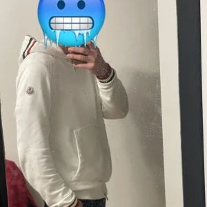 Vit Moncler hoodie med dragkedja - Säljer en clean vit hoodie från Moncler med dragkedja och huva. Tröjan har Moncler-logga på ärmen, snörning i huvan och snygga röd-blå-vita ränder på muddar och huva. Perfekt för dig som gillar stilrena och exklusiva plagg.