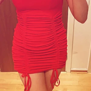 Röd tight kortklänning med snörning - Säljer en röd, figurnära kortklänning med snygga rynkade detaljer och snörning längs sidorna. Klänningen har korta ärmar och är tillverkad i ett stretchigt material som sitter skönt och framhäver formerna. Perfekt för dig som vill sticka ut med en färgstark look.