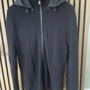 Mörkblå cardigan från Herno - Säljer en mörkblå cardigan från Herno med dragkedja framtill och huva i ett glansigt material. Tröjan har långa ärmar och ribbade muddar. Perfekt för dig som gillar stilren och bekväm design. Använd max 5 gånger och är i storlek 46(M)