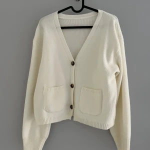 Vit croppad kofta med fickor - Säljer en vit croppad kofta med v-ringning och tre bruna knappar framtill. Koftan har två praktiska fickor och är stickad i mjukt material. Perfekt för lager-på-lager och har långa ärmar med breda muddar. Enkel och stilren design som passar till jeans eller kjol.