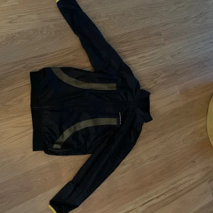 Svart Porsche vindjacka med gula detaljer - SUPERSNYGG PORSCHEJACKA! TRILL FÖR LAGRING NY FRÅN MIN PAPPASvart vindjacka från Porsche med snygga gula detaljer och logga på bröstet. Jackan har hel dragkedja framtill, meshfoder och sportig look. Perfekt för dig som gillar stilren design och vill sticka ut med exklusivt märke.