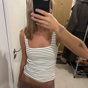 Randig ribbad linnetopp i vitt - Säljer en vit ribbad linnetopp med svarta tunna ränder och fyrkantig ringning. Toppen är ärmlös och har en tight passform som sitter snyggt på kroppen. Perfekt för varma dagar eller att styla lager på lager.