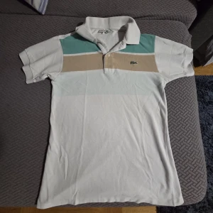 Randig pikétröja från Lacoste - Snygg pikétröja från Lacoste med breda ränder i vitt, beige och mintgrönt. Klassisk krage, korta ärmar och den ikoniska krokodilloggan på bröstet. Perfekt för en avslappnad och stilren look. Äkta, i nyskick. Använd endast någon gång, helt ren och fräsch utan fläckar.