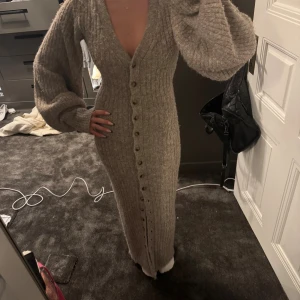 Beige stickad långklänning med knappar - Supermysig beige stickad långklänning med djup v-ringning och långa puffärmar. Klänningen har ribbad struktur och knappar hela vägen framtill, vilket ger en snygg och trendig look. Perfekt för dig som gillar att sticka ut med stil och komfort.