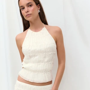 Vit linnetopp med snörning och öppen rygg - Säljer en vit  linnetopp med halterneck och snörning i ryggen. Toppen har ett plisserat tyg med tunna volangdetaljer i horisontella rader. Modellen är ärmlös och har öppen rygg, perfekt för varma dagar. Snygg och trendig design som ger en fräsch look.