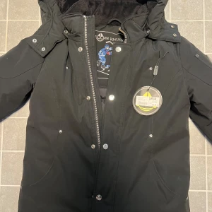 Svart parka från Moose Knuckles - Svart parka från Moose Knuckles med broderad hockeyspelare på insidan. Jackan har huva, dragkedja och tryckknappar framtill samt flera fickor. Fodrad för extra värme och med snygga detaljer i silver. Perfekt för kalla dagar.