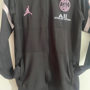 Svart PSG  - Svart träningsjacka från PSG x Jordan med rosa detaljer och loggor på bröstet. Jackan har hel dragkedja, fickor på sidorna och diskreta grå inslag på ärmarna. Perfekt för dig som gillar sportig och stilren streetwear.
