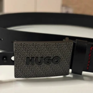 Svart skärp från Hugo Boss med röd söm - Snyggt svart bälte från Hugo Boss med bred, präglad metallspänne där det står HUGO. Bältet är i skinn och har coola röda sömmar som kontrast. Perfekt accessoar för att lyfta din outfit med en stilren och modern touch.