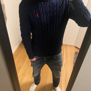 Mörkblå tröja Polo Ralph Lauren  - Säljer en mörkblå kabelstickad tröja från Polo Ralph Lauren med klassisk röd logga på bröstet. Storlek M. Jag är 76kg 186cm.