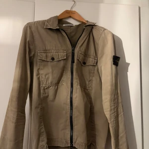 Beige overshirt från Stone Island - Säljer en beige overshirt från Stone Island med klassisk patch på ärmen. Jackan har två bröstfickor med knappar, dragkedja framtill och krage. Tillverkad i bomull och har en clean, stilren look som passar perfekt till streetwear.