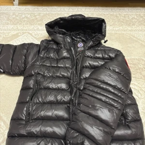 Svart dunjacka från Canada Goose S - Svart pufferjacka från Canada Goose i storlek S. Jackan har quiltad design, huva och dragkedjor framtill samt på fickorna. Klassisk logga på ärmen och glansig yta. Perfekt för kalla dagar när du vill hålla stilen och värmen.