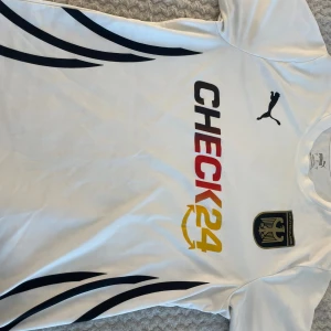 Vit fotbollströja från Puma Tyskland - Vit fotbollströja från Puma med Tysklands klubbmärke på bröstet och CHECK24-logga i svart, rött och gult. Snygga svarta detaljer längs sidorna. Tillverkad i lätt och ventilerande material, perfekt för träning eller match.