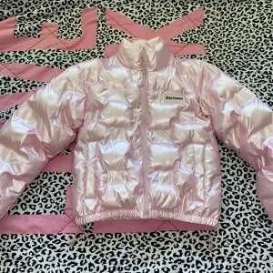 Rosa pufferjacka Juicy Couture S - Glansig rosa pufferjacka från Juicy Couture med broderad logga på bröstet och präglade JC-detaljer över hela jackan. Jackan har hög krage, dragkedja framtill och elastisk nederkant. Perfekt statement-plagg för dig som gillar att sticka ut.