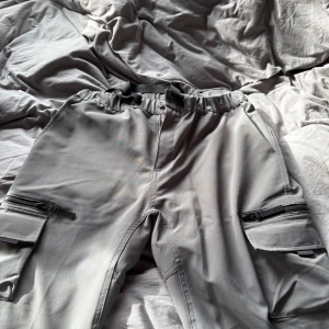Grå cargopants från Carhartt WIP - Säljer ett par grå cargopants från Carhartt WIP med elastisk midja och justerbart band. Byxorna har flera fickor med lock och dragkedja på sidorna, samt Carhartt-logga bak. Perfekta för en chill och streetig stil.