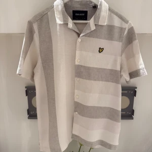 Randig skjorta från Lyle & Scott S - Kortärmad skjorta från Lyle & Scott i beige och gråa breda ränder. Klassisk krage, knäppning framtill och broderad gul logga på bröstet. Skön och luftig i bomull, perfekt för varma dagar.