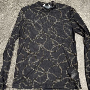 Svart mesh topp med kedjemönster - Snygg svart mesh topp från C i storlek XS. Toppen har lång ärm och är täckt av ett coolt kedjemönster i guld. Materialet är tunt och stretchigt, perfekt för att skapa en trendig lager-på-lager-look. Hög hals ger en extra stilren vibe.