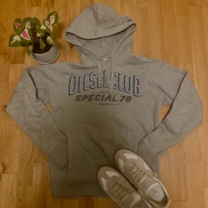 Diesel Hoodie - Snygg grå hoodie från Diesel med blått tryck på bröstet där det står 'Diesel Club Special 78'. Tröjan har huva med snörning och en klassisk känguruficka framtill. Perfekt för en chill och avslappnad stil. Riktigt bra kvalitet inga skador eller fläckar, sitter mer som S inte L