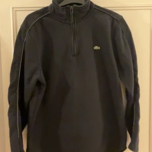 Svart fleece half zip från Lacoste - Svart fleece-tröja från Lacoste Sport med half zip och hög krage. Klassisk krokodil-logga på bröstet och diskreta sömdetaljer längs axlarna. Perfekt för kyliga dagar och sportiga outfits.