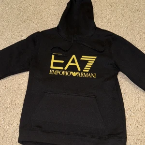 Svart EA7 Emporio Armani hoodie M - Svart hoodie från EA7 Emporio Armani med stor gul logga på bröstet och snyggt tryck på ryggen. Tröjan har huva med snörning och en klassisk magficka. Materialet är mjukt och känns skönt mot huden, perfekt för chill eller streetwear.