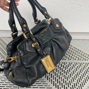 Svart Marc by Marc Jacobs handväska - Snygg svart handväska från Marc by Marc Jacobs i äkta skinn med guldfärgade metalldetaljer och dragkedja. Väskan har dubbla handtag och en cool logga i metall framtill. Fodret är mönstrat med stora bokstäver i svart och vitt. My lowewst price 🙏❤️