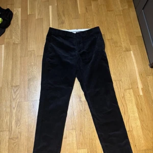 Svarta manchesterbyxor från Gant - Svarta manchesterbyxor från Gant i klassisk rak modell. Byxorna har diskreta fickor fram och bak, och är tillverkade i mjuk manchester som ger en snygg och stilren look. Perfekta för dig som gillar en tidlös och clean stil. Storleken 170/82 på byxor ungefär 170 cm lång och har ett midjemått på 82 cm.