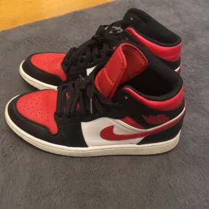 Nike Air Jordan 1 svart/röd/vit - Säljer ett par Nike Air Jordan 1 sneakers i klassisk svart, röd och vit färg. Skorna har högt skaft, svart snörning och den ikoniska röda swoosh-loggan på sidan. Ovandelen är i skinn och de har en vit mellansula med svart yttersula. Perfekta för dig som gillar streetstyle. Boxen följer med