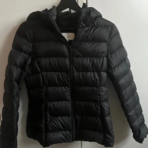 Svart pufferjacka från Vila med huva - Säljer en svart pufferjacka från Vila med huva och dragkedja framtill. Jackan är quiltad med horisontella sömmar och har en figurnära passform. Perfekt för kyliga dagar och enkel att matcha med olika outfits.