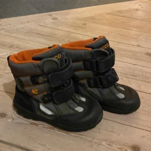 Svarta vinterkängor från Ecco - Säljer ett par svarta och grå vinterkängor från Ecco med orange foder. Skorna har två breda kardborreband för enkel påtagning och robust platt sula. Materialmix av syntet och skinn, perfekta för kalla dagar.storlek 28, hör av dig om du har frågor eller funderingar!