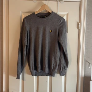 Grå tröja från Lyle & Scott - Snygg grå tröja från Lyle & Scott med klassisk rund halsringning och gul broderad logga på bröstet. Tröjan är långärmad och har ribbade muddar vid ärmslut och nederkant. Perfekt för lager-på-lager under hösten och vintern.