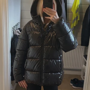 Svart glansig pufferjacka med huva - Säljer en svart glansig pufferjacka med huva och dragkedja framtill. Jackan är vadderad och har en oversized look, perfekt för kalla dagar. Den har långa ärmar och breda kanaler som ger en cool och modern vibe.