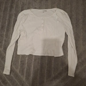 Vit ribbad långärmad tröja H&M Divided - Vit långärmad tröja från H&M Divided med ribbad struktur och rund halsringning. Tröjan har knappar framtill och är i en croppad modell som ger en avslappnad vibe. Perfekt att matcha med jeans eller kjol för en enkel och clean look.