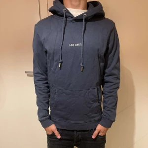 Mörkblå hoodie från Les Deux - Säljer en mörkblå hoodie från Les Deux med vit broderad logga på bröstet. Tröjan har huva med snörning, känguruficka och ribbade muddar. Perfekt för dig som gillar en clean och stilren look.