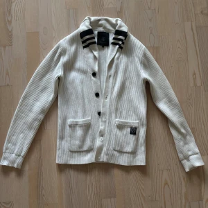 Vit Cardigan från Jack & Jones Premium - Ribbstickad vit kofta från Jack & Jones Premium med svarta ränder på kragen, svarta knappar och två fickor framtill. Klassisk och stilren design som passar perfekt till lager-på-lager. Mjuk och skön känsla.