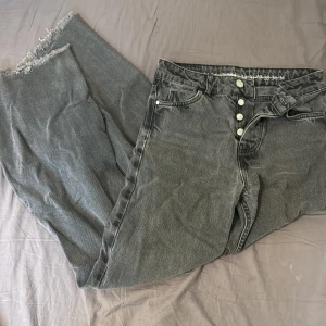 Gråa raka jeans med råa kanter high waist - Säljer ett par grå jeans med raka ben och råa, fransiga benslut. Jeansen har knäppning med flera silverfärgade knappar och klassisk femficksdesign. Snygg, lite tvättad look och passar perfekt till en avslappnad stil.