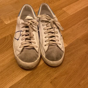 Philippe Model vita sneakers med blå detalj - Säljer ett par vita Philippe Model sneakers i skinn med grå mockadetaljer på tån, blå hälkappa och röd kant. Snygg blå logga på sidan och klassisk snörning. Tillverkade i Italien och riktigt stilrena till jeans eller shorts.